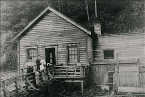 A Japanese man and woman outside a small wooden house. The man is holding a baby. There is a baby carriage on the porch. 